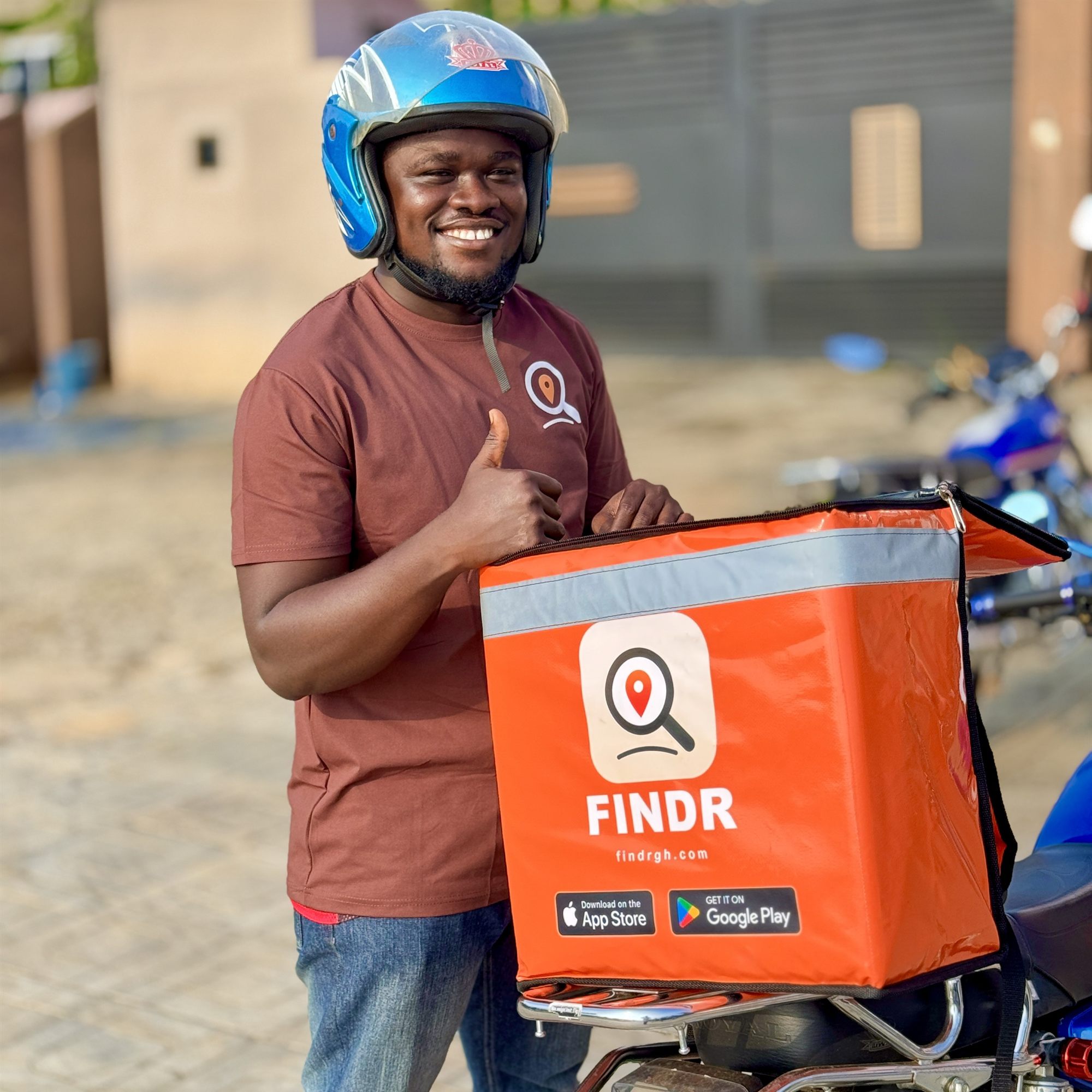 A smiling FINDR delivery rider in a blue helmet giving a thumbs up beside his orange FINDR delivery box